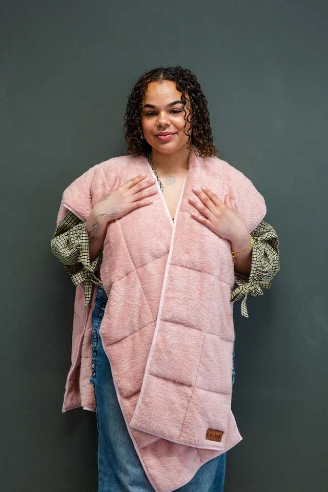 A person with curly hair wearing a plaid shirt and jeans is draped in a pink CALMR Shawl, standing against a plain dark gray background.
